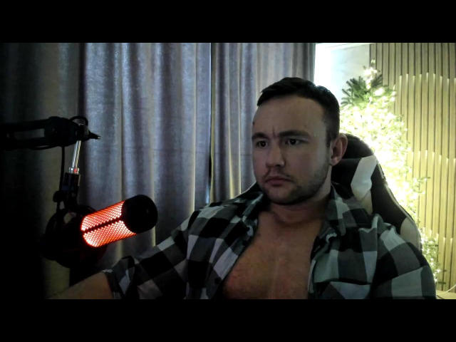 MarkusFit with a serious face, shirt unbuttoned, chest visible, sitting in a chair, dark hair, camera on left, red light, black and white checkered shirt, green curtains.