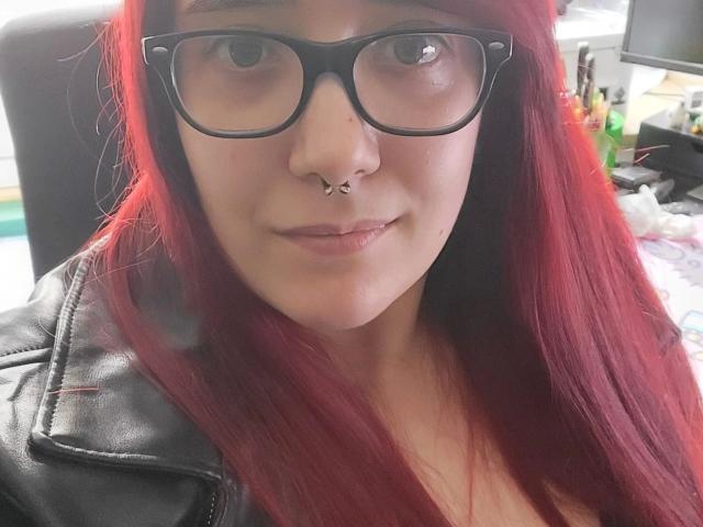LadyJosefine-hot with red hair, black glasses, and a nose piercing, wearing a black leather jacket, smiling slightly. Her tits are partially visible, and her pussy is not.