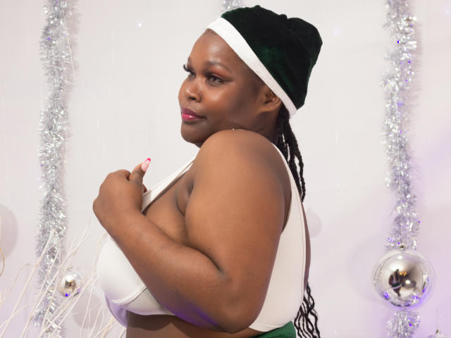 SheilaTorrez, com pele escura, usando um gorro de Papai Noel verde de veludo, um sutiã branco, mostrando seus seios grandes, redondos e firmes, olhando por cima do ombro, com batom rosa e unhas longas..