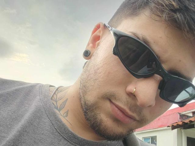 Sthefano with a beard, black sunglasses, nose ring, and ear gauge, wearing a gray shirt, showing a tattoo on his neck, under a cloudy sky.