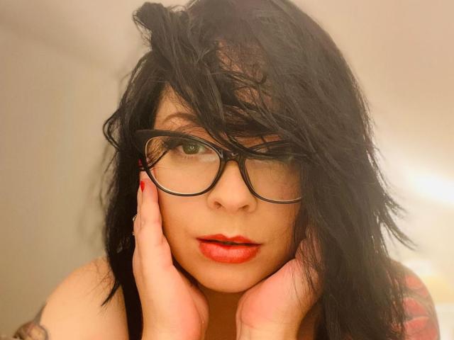 RenataEvergreen has black messy hair, green eyes, red lipstick, and black glasses. She's touching her face with red-manicured fingers, looking slightly confused. Her tits are partially visible.