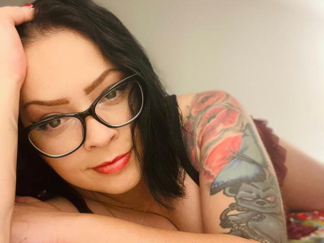 RenataEvergreen has black glasses, red lipstick, and a tattooed arm with roses and a skull on her right side, lying down with a hand in her black hair.