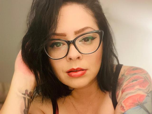 RenataEvergreen with black glasses, green eye makeup, red lipstick, black hair, tattooed arm, and black top, looking directly at camera. Boobs visible in top. Soft light.