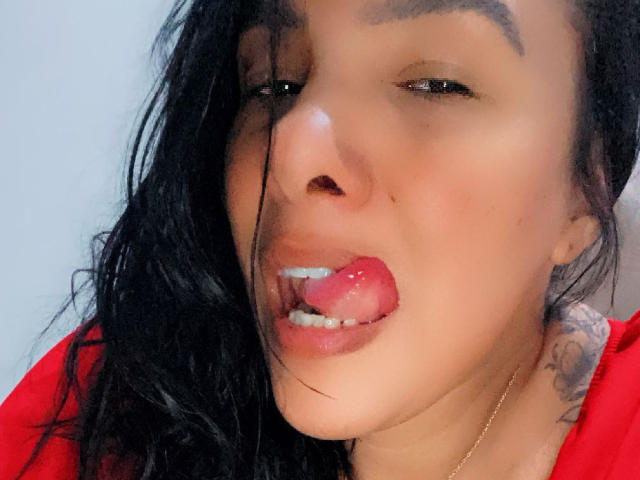 RondaHot's face with wet black hair, brown eyes, and red shirt, tongue out, showing a small amount of white teeth, and a tattoo on the right side of her neck.
