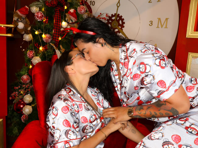 DilanAndMia con cabello negro y una diadema roja besando a DilanAndMia con cabello castaño, vistiendo pijamas blancos a juego con caras de Santa, tomados de la mano, en un ambiente navideño acogedor..
