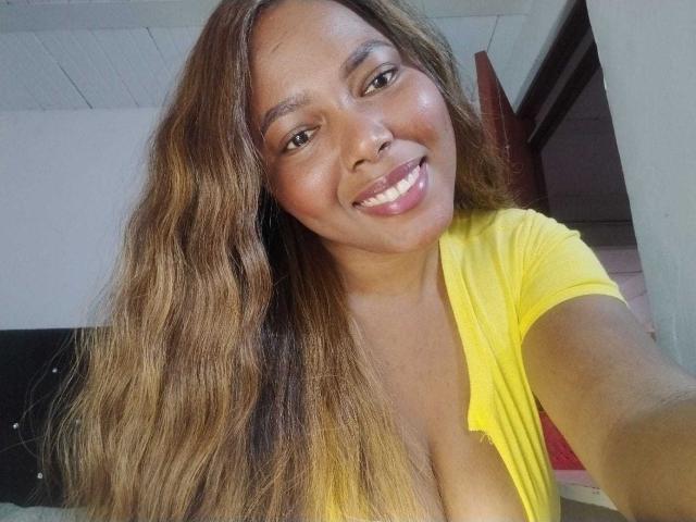 EmiraHot with long brown wavy hair, smiling, wearing a low-cut yellow shirt, showing her tits, in a selfie, has a cute nose ring and shiny pink lipstick.