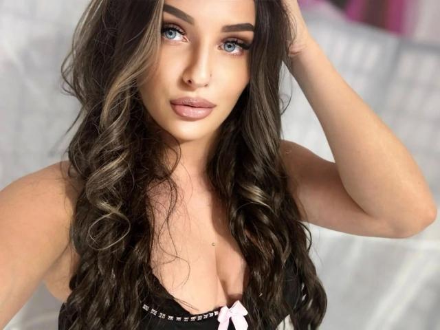 SweetSerena-hot with long, wavy brown hair, blue eyes, and pink lips, wearing a black bra with a pink bow, touching her hair. Boobs are visible, cleavage highlighted.