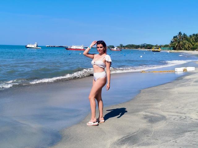 ZoeeMar stands on a beach in a white bikini, hand in hair, showing her tits and ass. She's wearing white sandals, with boats in the blue sea.