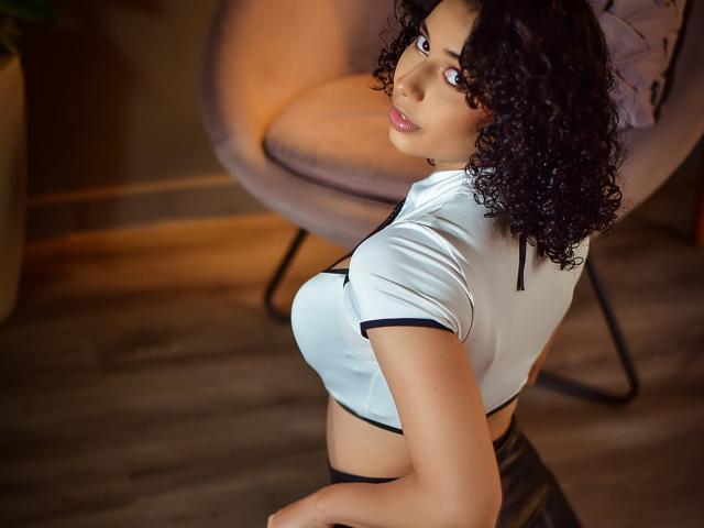 Zelphirina, con cabello negro y rizado, mirando por encima del hombro, vestida con una blusa blanca ajustada que deja ver sus grandes senos, y con ropa interior negra, en una habitación con luz tenue..