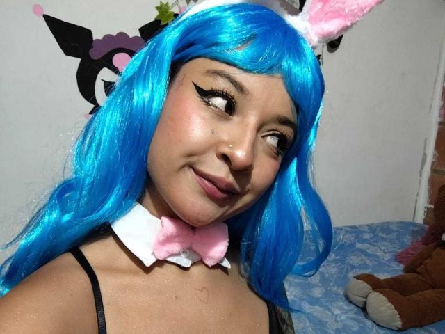 JuanaAhh con peluca azul, lazo rosa, orejas de conejo blancas y delineador de ojos negro, sonriendo de lado, mostrando su piel brillante y tersa, y un pequeño tatuaje de corazón en su pecho..