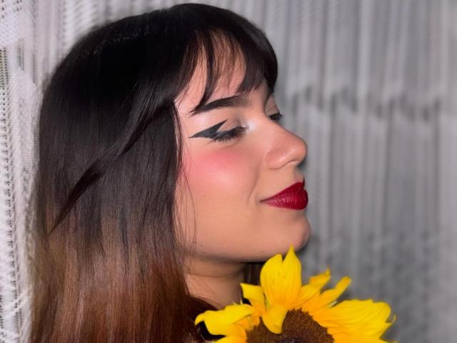 Milly Johnson, com cabelo castanho escuro, batom vermelho e delineador preto marcante, segurando um girassol, sorrindo com os olhos fechados. Suas nádegas não estão visíveis..