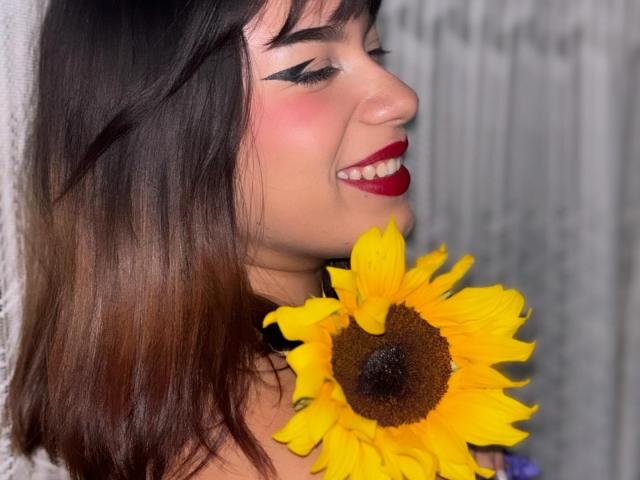 Milly Johnson, com cabelo castanho escuro, batom vermelho, delineador preto, sorrindo, segurando um girassol amarelo vibrante contra uma parede branca. Seus seios não estão visíveis..