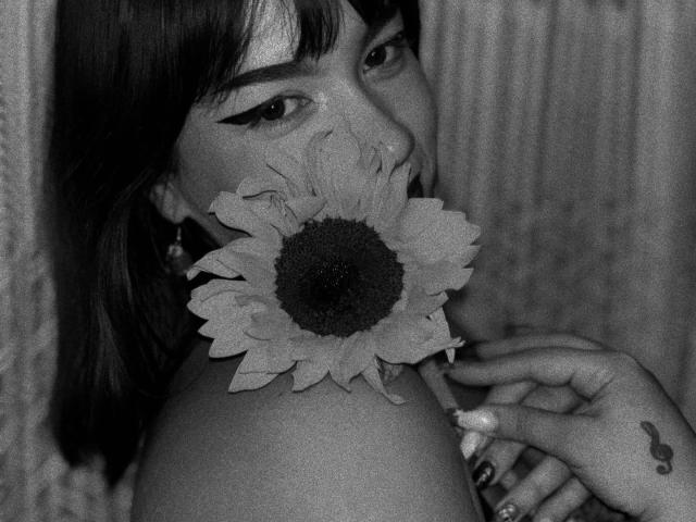 Fotografía en blanco y negro de Milly Johnson con un girasol cubriendo sus pechos, mirando por encima del hombro, delineador de ojos "alas de la paz", cabello oscuro, tatuaje discreto en la mano derecha..