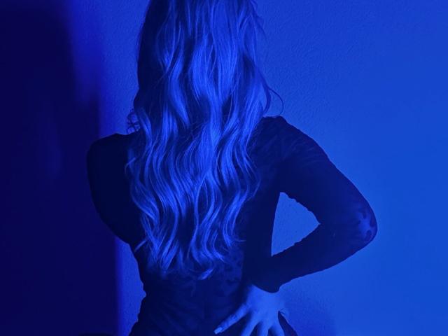 BalletSwan with long wavy blonde hair, facing away, hands on hips, wearing a black sheer long-sleeve top, illuminated by blue light.