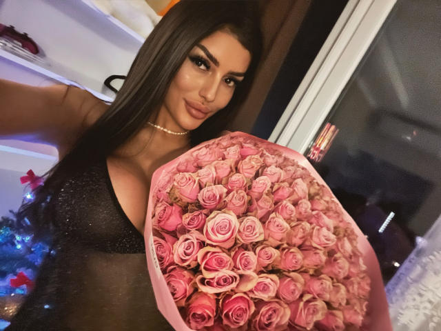 ChocoLatess with long black hair, wearing a sparkly black dress, holding a bouquet of pink roses, showing her big tits, in a dimly lit room.