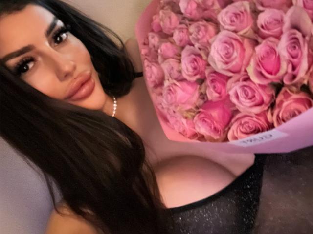 ChocoLatess with long black hair, full lips, and large tits, wearing a black top, holding a pink rose bouquet. She's wearing a pearl necklace.