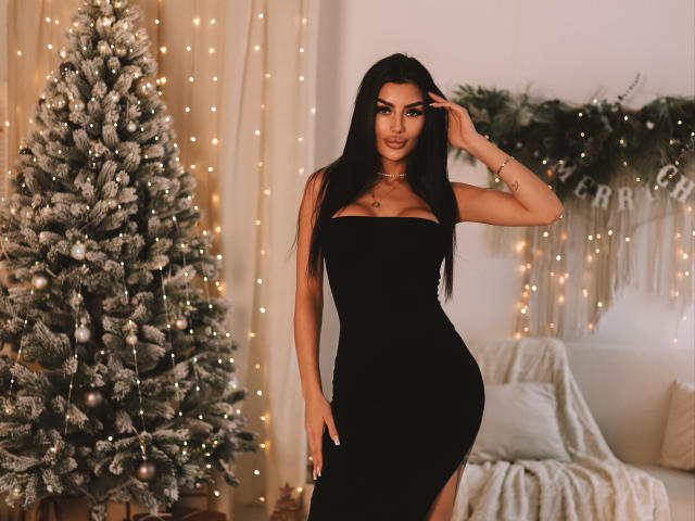 ChocoLatess with long black hair, wearing a tight black dress, standing next to a frosted Christmas tree, hand on her head, glowing fairy lights in the background.