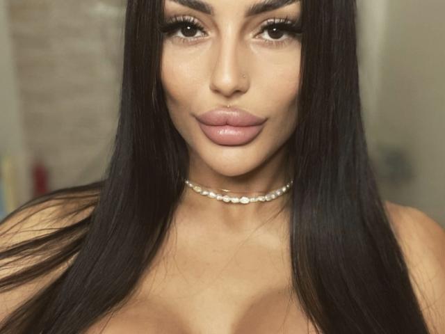 ChocoLatess has long black hair, full pink lips, a shiny nose, and a pearl choker, showing off her tits and a sexy smile in this shot.