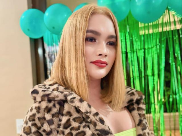 HornyCumTs with blonde bob, wearing leopard print coat, green strap dress, red lipstick, and green balloons in background, looking cute and confident. Boobs slightly visible under coat.