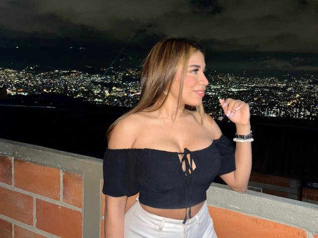 LunaMiranda with medium tits in a black off-shoulder top, white high-waisted pants, holding a cigarette, night sky, city lights in the background, brick balcony, smiling, right profile.