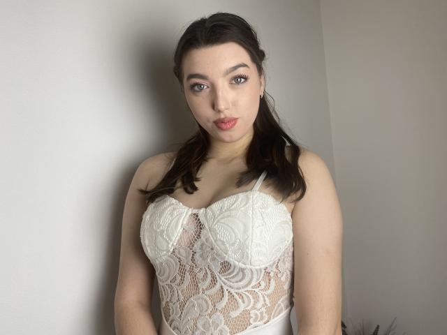 Vailett with dark hair, wearing a white lace bodice, showing her tits, pale skin, and red lipstick, standing against a plain wall, looking at the camera.