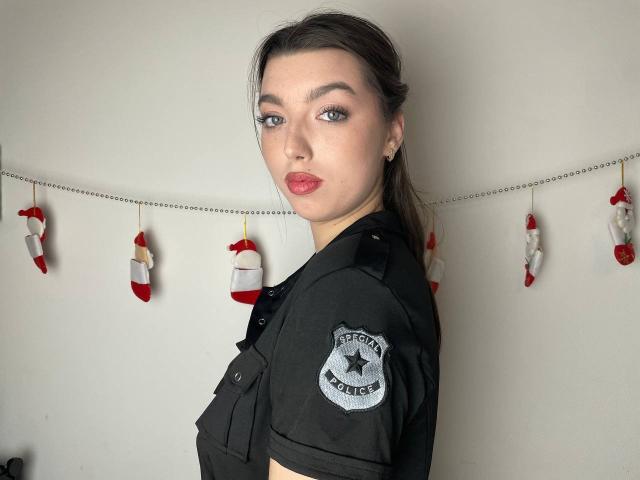 Vailett with pouty red lips, wearing black police uniform, showing her shoulder, brown hair in a ponytail, blue eyes, and a police badge on her sleeve.