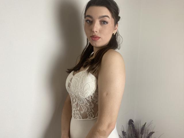 Vailett, avec une peau pâle, des cheveux bruns foncés et du rouge à lèvres, portant un corsage blanc en dentelle, regardant la caméra, les bras baissés, de petites boucles d'oreilles, une ombre sur un mur blanc..
