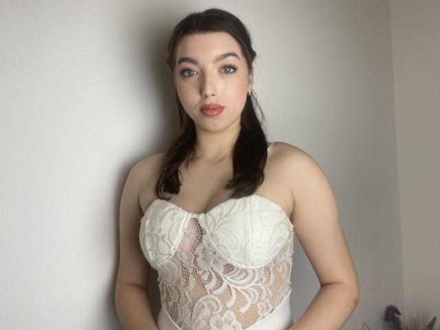 Vailett, con piel pálida, cabello castaño oscuro en trenzas, vestida con un vestido blanco de encaje sin tirantes, mostrando sus senos grandes, firmes y ligeramente redondeados contra una pared lisa..