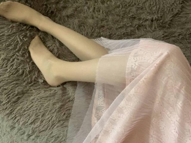 Lialit's legs are crossed on a grey, fluffy surface, wearing a pink, lace-patterned dress with a sheer, white overlay. Her pale, nude stockings highlight her slender, fair-skinned legs.