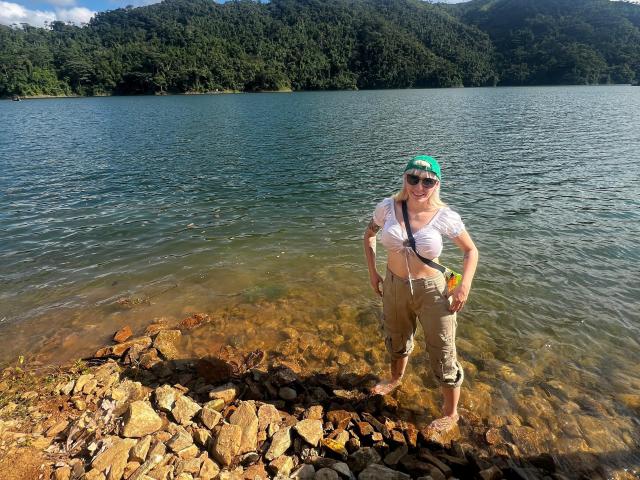 SaraleePoison stands in a rocky, shallow lake, wearing a green cap, white shirt, and khaki pants, with a black strap across her tits, looking at the camera.