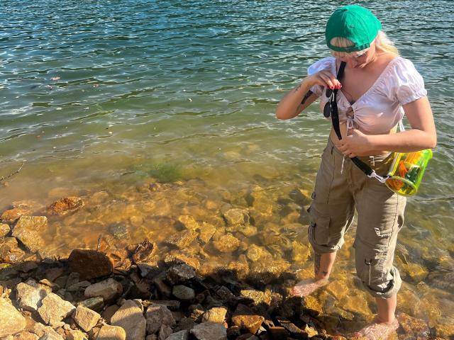 SaraleePoison stands in clear water, wearing a green cap, white shirt tied at the chest, and khaki pants. She holds a camera and a yellow bag.