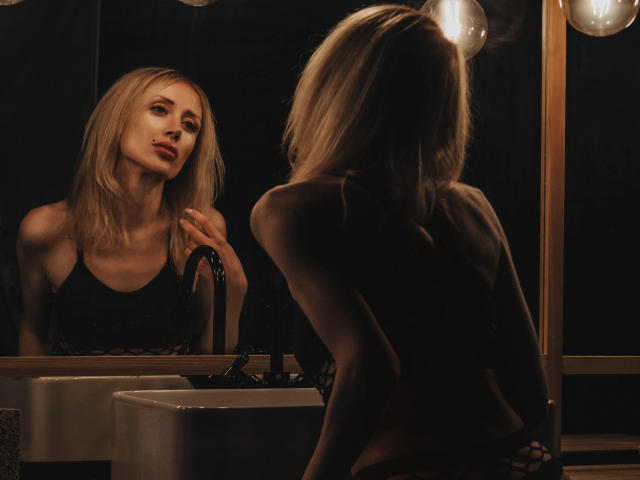 ReginaJune with blonde hair, looking at herself in a mirror, wearing a black top, standing in front of a sink, reflecting in a dimly lit room.