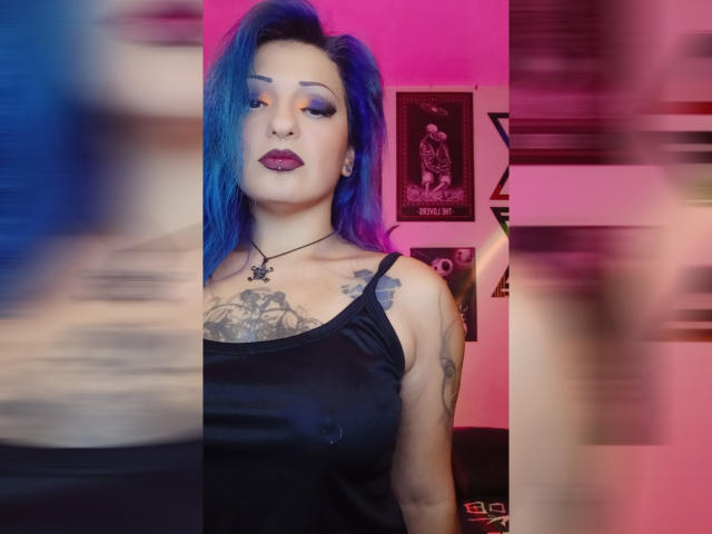 AnnyFreyr mit blau-lila Haaren, dunklem Lippenstift, einem schwarzen Tanktop, das Brüste und Tattoos zeigt, und einer Kreuzkette um den Hals, in einem verschwommenen, rosa und lila beleuchteten Ambiente..