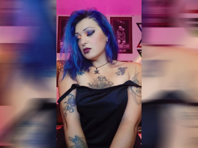 AnnyFreyr, avec ses cheveux bleus, ses tatouages et son maquillage foncé, porte un haut noir à une épaule qui dévoile sa poitrine, et un collier en forme d'étoile, le regard baissé avec une expression suggestive..