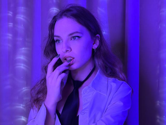 MissDi with purple-lit face, wavy brown hair, blue eyes, black choker, white shirt, and black top, sucking her finger, showing her pouty lips and small nose.