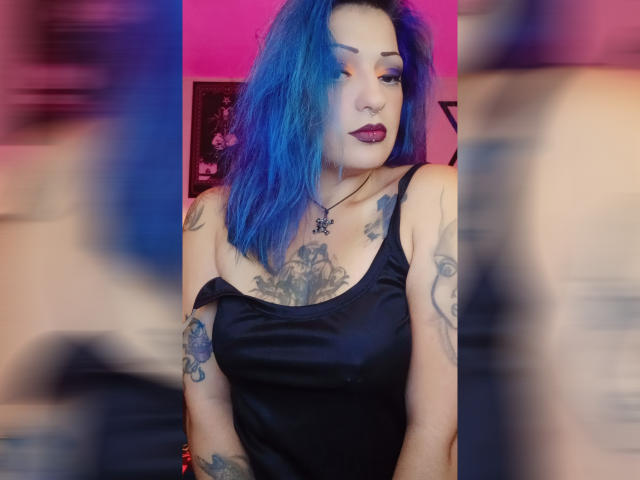 AnnyFreyr, com cabelo azul, vestido de cetim preto, tatuagens no peito e nos braços, batom escuro e um colar com uma cruz, olhando para a câmera, com reflexos borrados nas laterais..