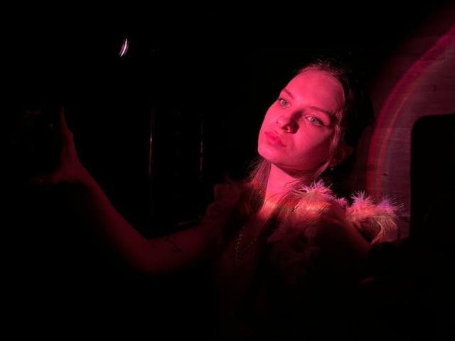 MissDi with pinkish-red lighting, serious expression, arms raised, wearing fluffy shoulder accessory, dark background, subtle tattoo on right arm, slight blush, looking slightly up.