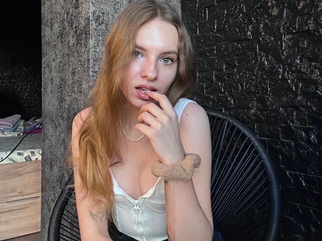MissDi with long blonde hair, white tank top, showing her tits, finger in mouth, scrunchie on wrist, pearl necklace, sitting in black chair, black textured wall behind.