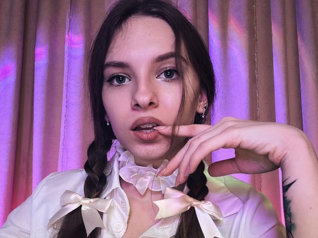MissDi with pouty lips, finger in mouth, wearing white dress with ribbons, braided hair, pink and purple light, visible tattoo on right wrist.