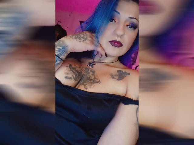 AnnyFreyr con i capelli blu, tatuaggi sul petto e sul braccio, top nero con una spalla scoperta, rossetto viola e piercing al naso, con una mano sul mento, sfondo sfocato. Seno, tatuaggi, espressione sicura..