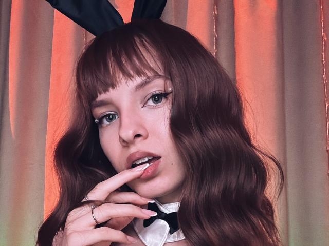 MissDi with wavy brown hair, black bunny ears, and a white collar, biting her finger with her pink lips and green eyes. Her ringed finger touches her mouth.