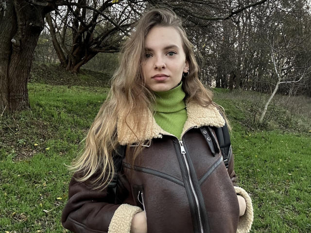 MissDi with long blonde hair, green turtleneck, and brown leather jacket with sheepskin lining, standing outdoors, looking serious, grassy field in background, overcast day.