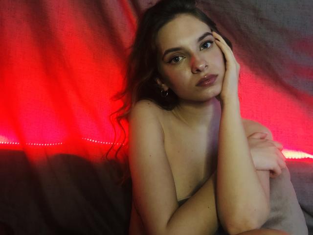 Nude MissDi with wavy brown hair, green eyes, and plump lips, holding her head with one hand, covering her tits with the other, under red lighting.