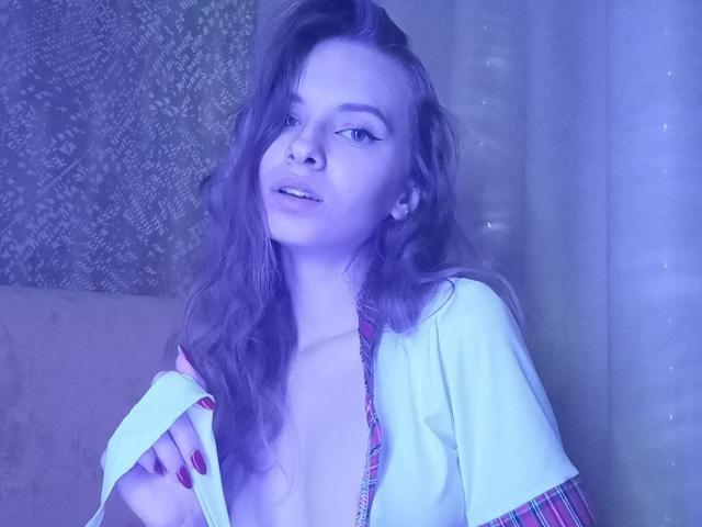 MissDi with wavy hair, light skin, and red nails, wearing a loose, partially open, colorful top, exposing her small, perky tits, in a blue-lit photo.