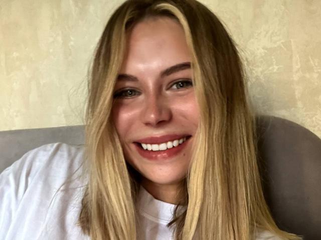 SassyJam has blonde hair, a white shirt, and a cute smile showing her pearly white teeth. She's looking at the camera with green eyes and a slight blush.