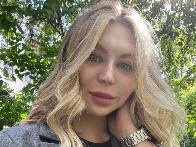 SassyJam, con cabello rubio ondulado, ojos verdes y lápiz labial rosa, vestida con un blazer gris, se toca el cuello, lleva un reloj dorado, y el fondo es un bosque soleado..