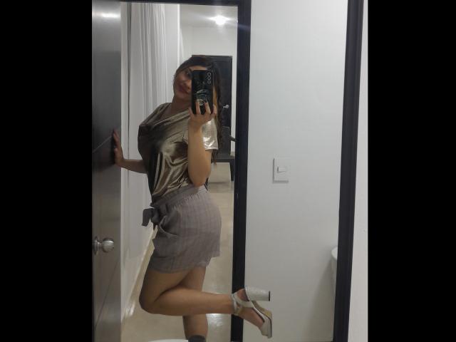 JulietaFoster69 in a shiny gold top and plaid mini-skirt, holding a phone, posing with one leg up, showing her ass and wearing white high heels.