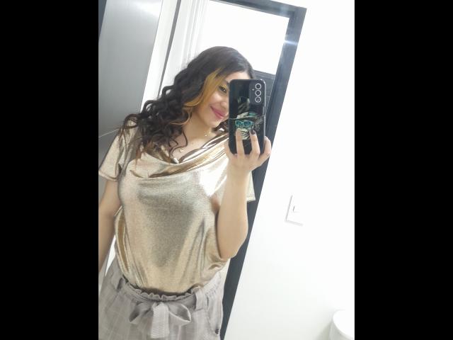 JulietaFoster69 with wavy brown hair, wearing a shiny gold top, showing her small tits, and grey tulle skirt, holding a phone with a black case, smiling.