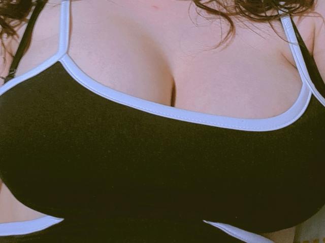JulietaFoster69's huge tits in a black sports bra with white trim, showing her ample cleavage and fair skin. Her brown curly hair is partially visible.