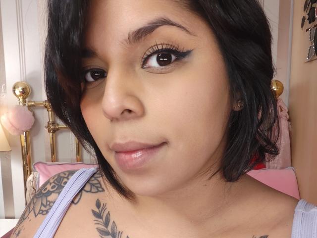 Chili with light brown skin and black hair is smiling with glossy lips, showing a tattoo on her right shoulder.